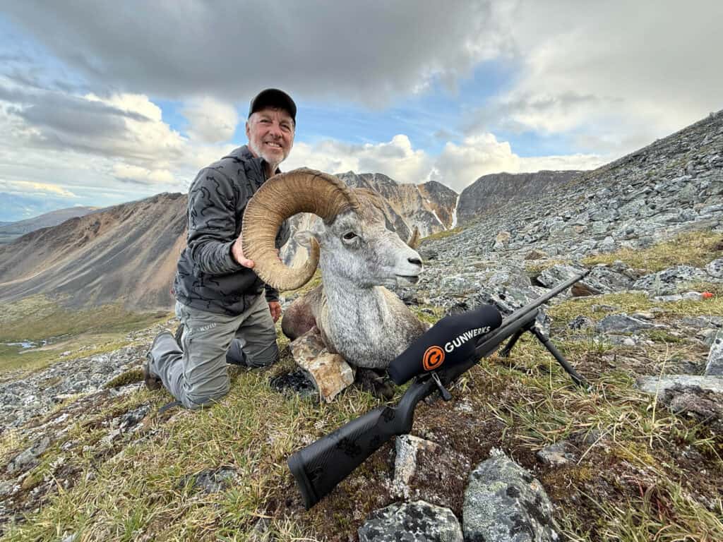 Hiker posing next to a beautiful mountain goat in rugged mountain terrain with a rifle.