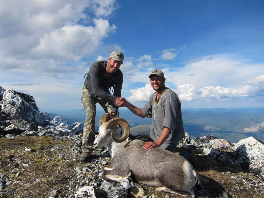 Two men shaking hands over a trophy elk on a mountain summit at Scoop Lake Outfitters.