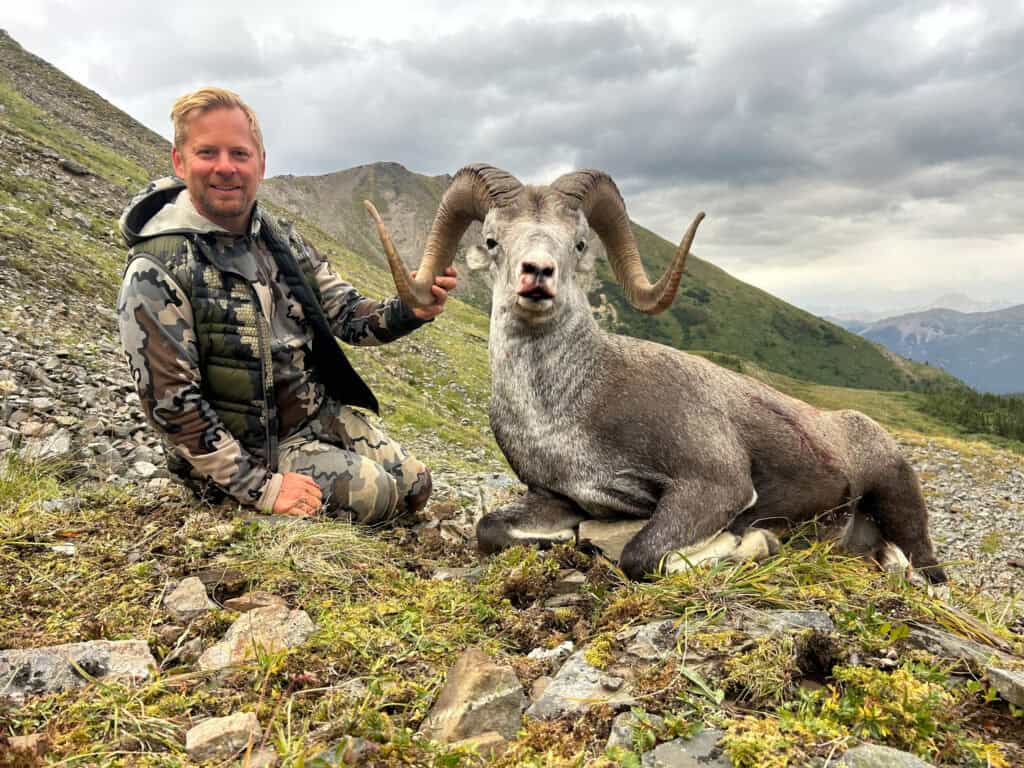 Hiker dressed in camouflage gear holding large horns of mountain goat in wild landscape.